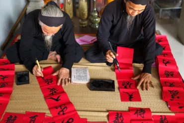 Two elderly men in black clothing write Chinese calligraphy on red banners laid out on a mat.