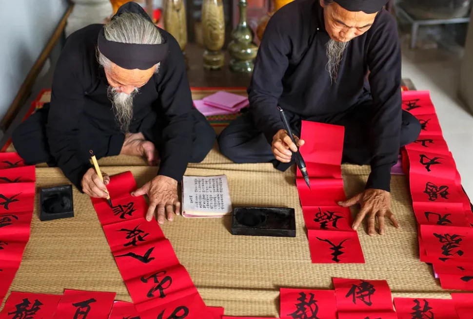 Two elderly men in black clothing write Chinese calligraphy on red banners laid out on a mat.