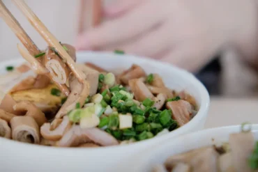 Chopsticks lift noodles from a bowl topped with chopped green onions; another bowl is out of focus in the foreground.