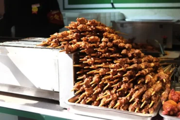 Pyramid of grilled meat skewers stacked on a metal tray at a street food stall.