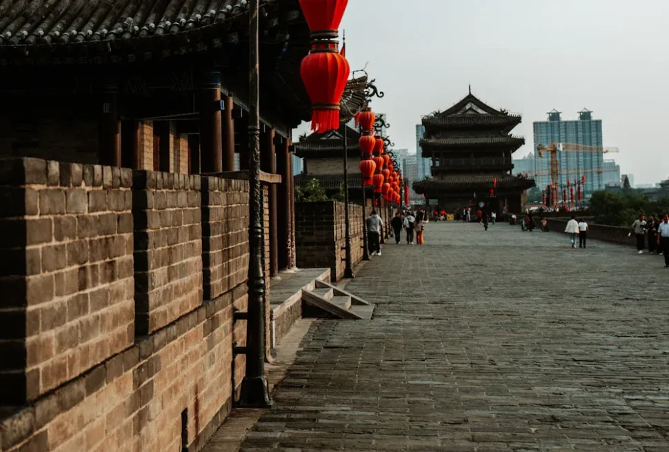 Stone wall and wide walkway along a historic Chinese street, red lanterns hang above, leading to a multi-tiered gate with modern buildings in the background.