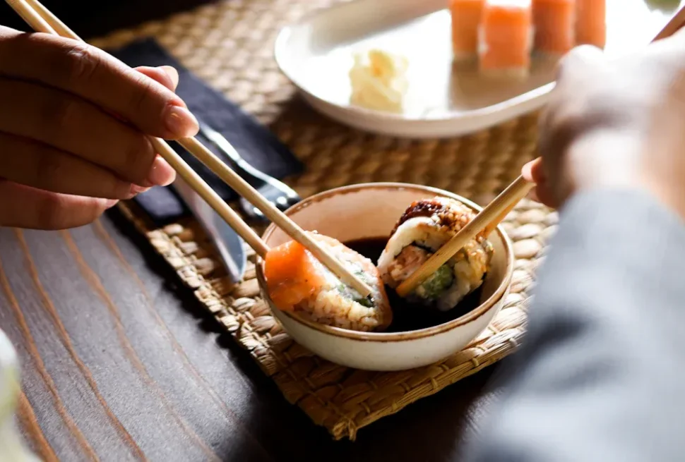 Hands use chopsticks to pick sushi from a small bowl on a woven placemat at a dining table.
