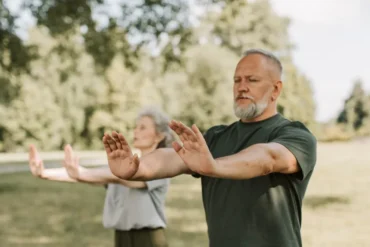 Two seniors practicing Tai Chi in a sunny park, arms extended forward in slow, controlled movements.