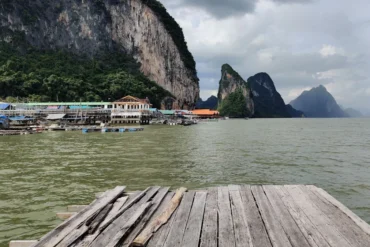Weathered wooden dock extends over calm water toward a coastal village and towering limestone cliffs.