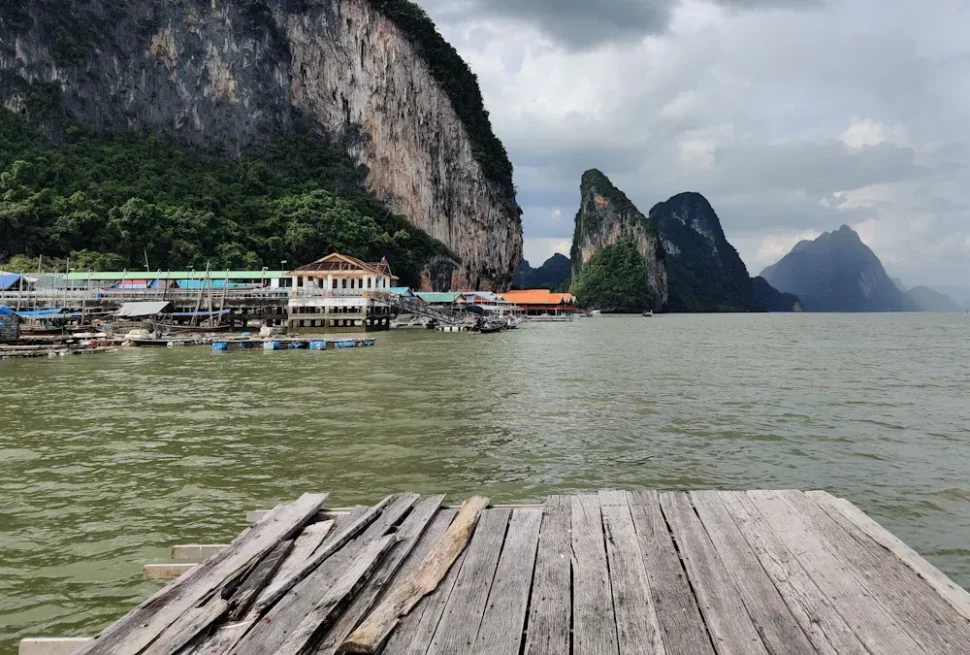 Yangshuo,Chinatour Weathered wooden dock extends over calm water toward a coastal village and towering limestone cliffs.