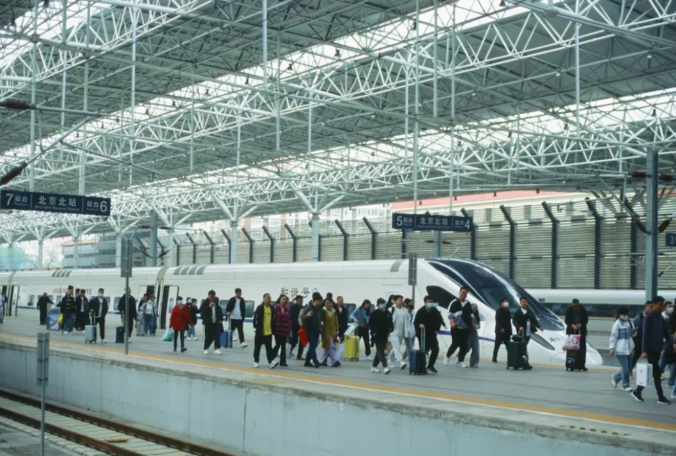 Busy train station platform with a white high-speed train, travelers carrying luggage and boarding the train under a steel lattice roof.