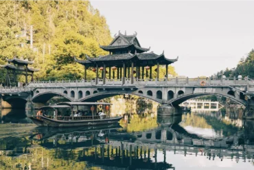 Stone arch bridge with a multi-tiered Chinese pavilion on top, spanning a calm river with a boat passing underneath and trees in the background in autumn light.