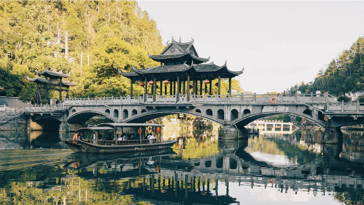 Stone arch bridge with a multi-tiered Chinese pavilion on top, spanning a calm river with a boat passing underneath and trees in the background in autumn light.