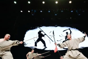 Three martial artists in beige robes wield wooden staffs and swords on a stage, dramatic action against a white brushstroke backdrop.