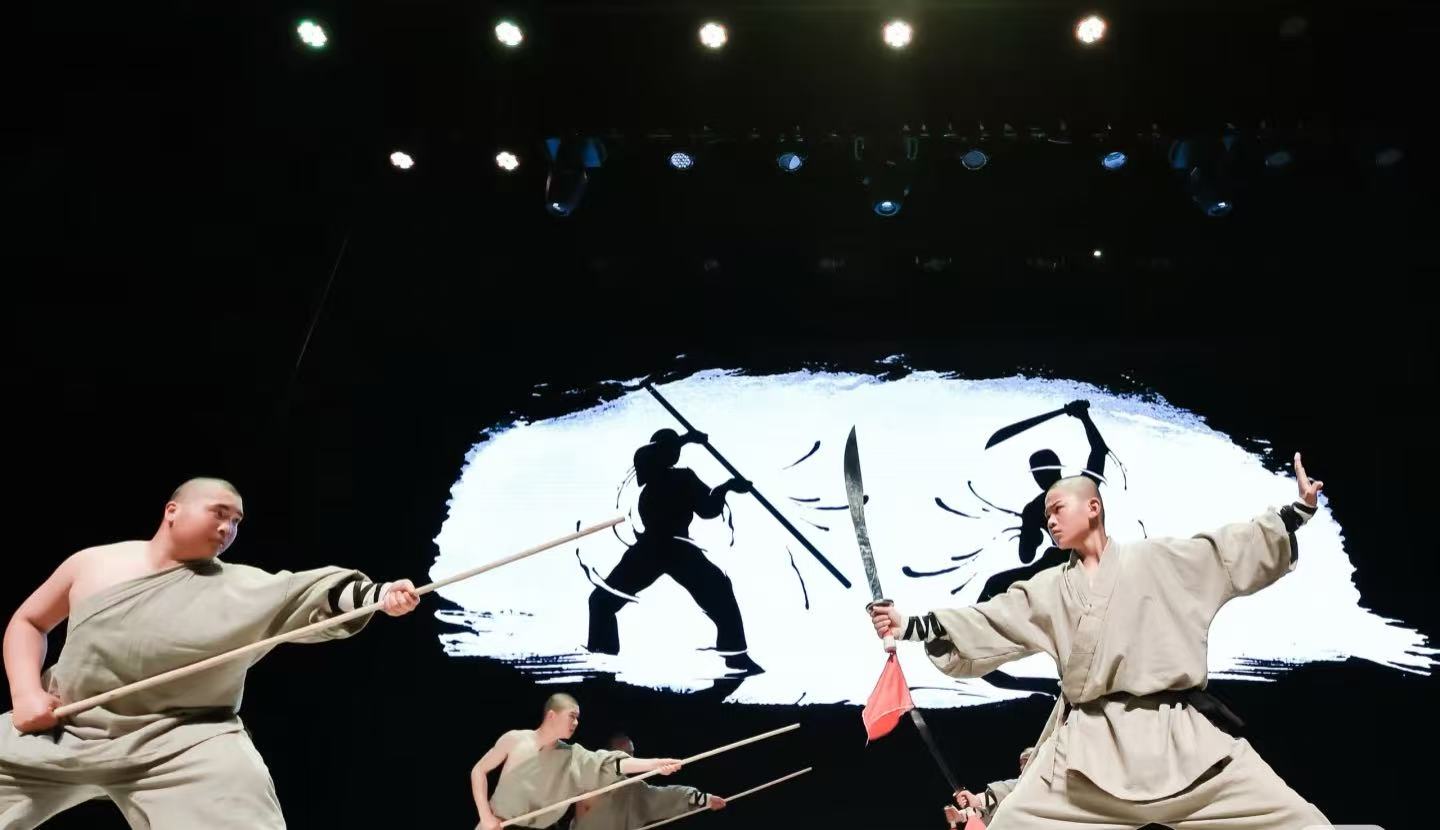 Beijing Kung Fu Show,Chinatour Three martial artists in beige robes wield wooden staffs and swords on a stage, dramatic action against a white brushstroke backdrop.