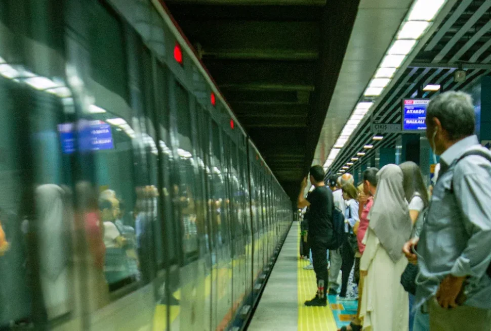 People wait on a subway platform beside a closing glass train car, with one rider wearing a hijab and others facing the train.