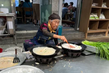 Elderly woman cooking crepe-like pancakes on two large griddles at a street stall, with fresh greens nearby.