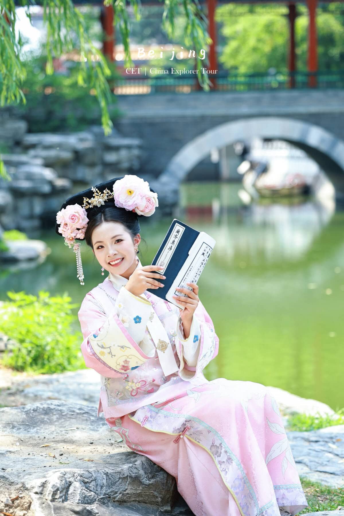 hanfu photoshoot, the forbidden city, beijing, qing dynasty, female, solo, chinatravel, Young woman in a pastel pink kimono with a floral headpiece sits by a pond, holding books near a stone bridge.