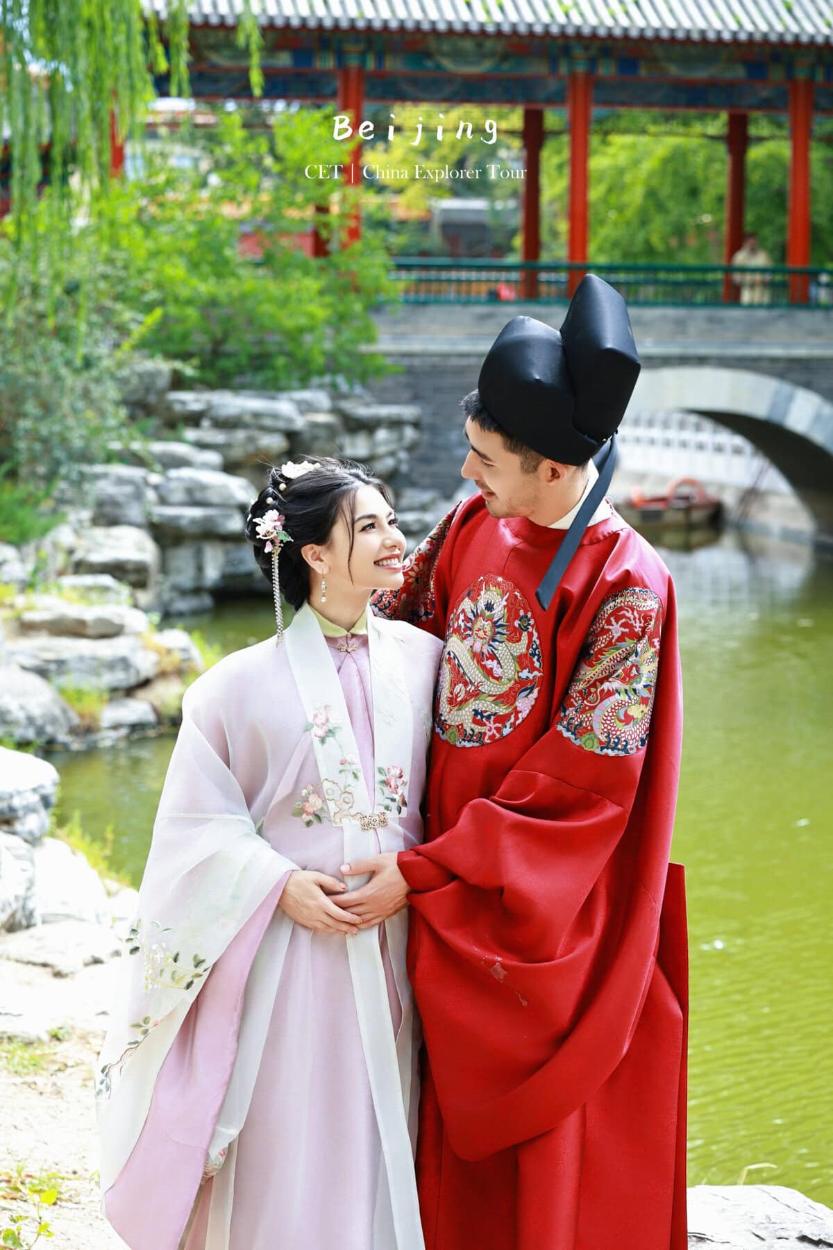 Hanfu photoshoot, Beijing, zhongshan park, couple, chinatravel, Couple in traditional Chinese wedding attire posing by a pond in a garden with a red pavilion in the background.