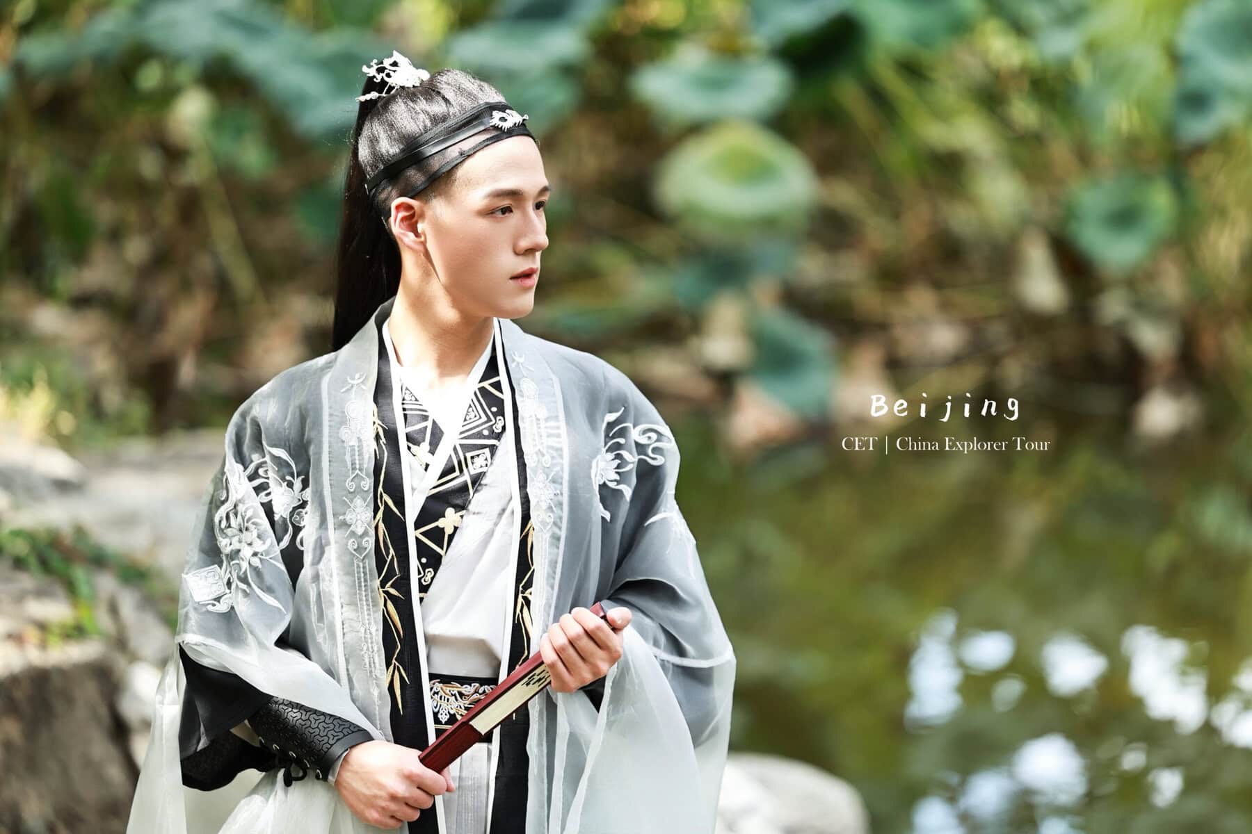 Hanfu Photoshoot, Beijing, zhongshan park, male, chinatravel, Person in traditional Chinese robe holding a wooden fan, standing outdoors by a water feature with green foliage; 'Beijing CET | China Explorer Tour' text visible on the right.