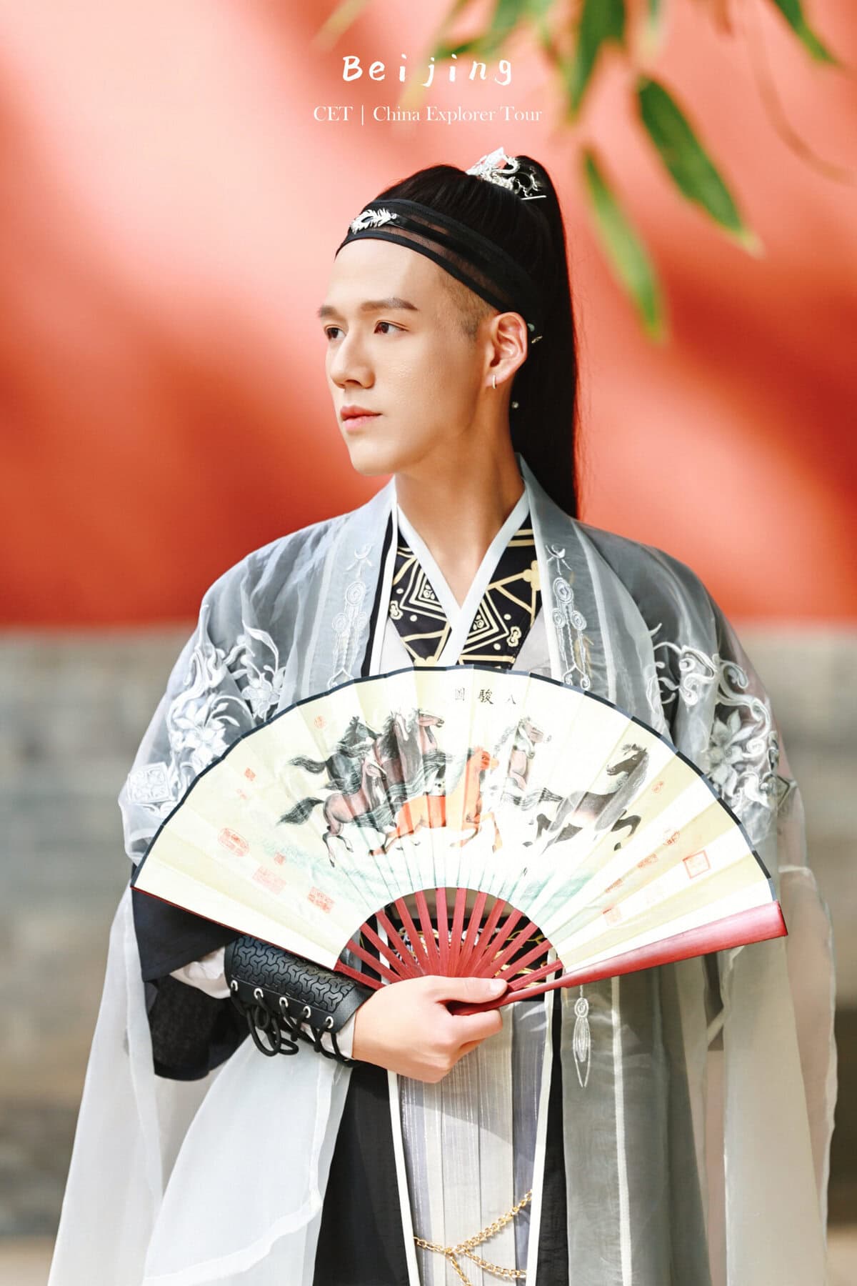 Hanfu Photoshoot, Beijing, zhongshan park, male, chinatravel, Person in traditional Chinese hanfu dress holding a decorative fan, standing against a red background with greenery above