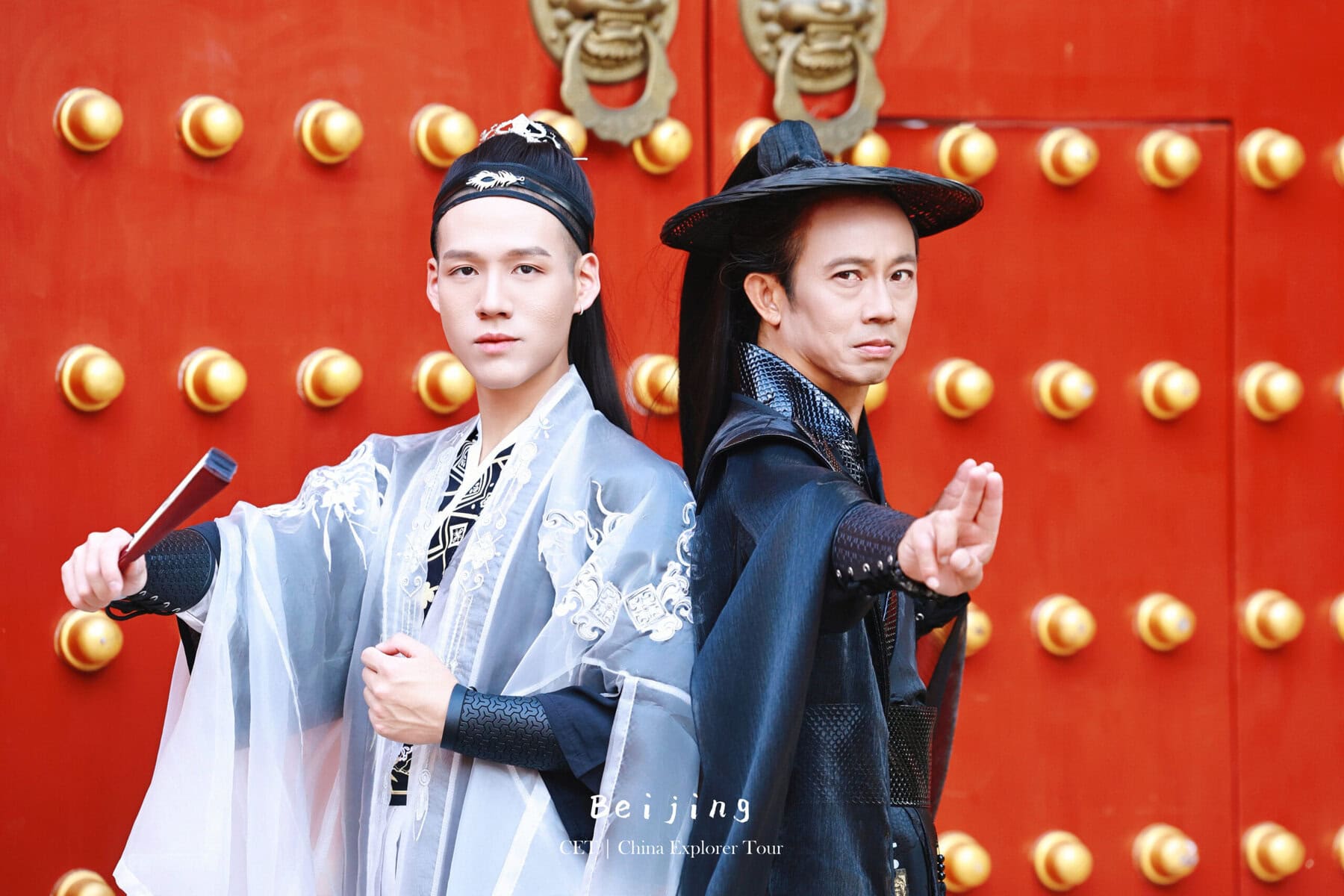 Hanfu Photoshoot, Beijing, forbidden city exterior, male, friends, chinatravel, Two performers in ornate traditional Chinese costumes pose back-to-back in front of a bright red, studded door; one in pale blue robes and the other in dark clothing, striking martial-arts stances. Text at bottom reads 'Beijing' and 'China Explorer Tour'.