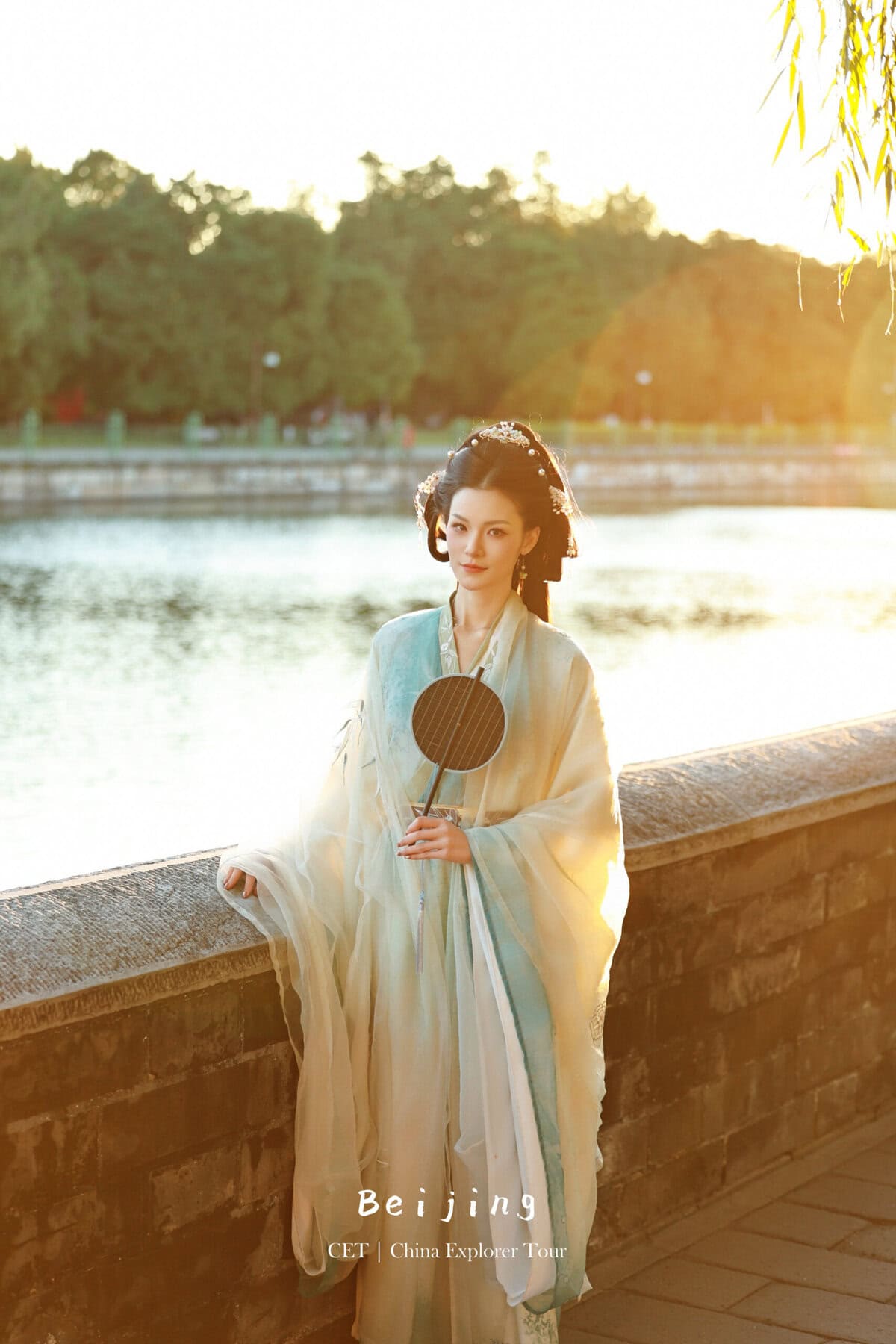 Hanfu photoshoot, Beijing, forbidden city exterior, female, chinatravel, Portrait of a woman in traditional Hanfu standing by a stone railing along a river at sunset, holding a round fan; text at bottom reads ‘Beijing CET | China Explorer Tour’