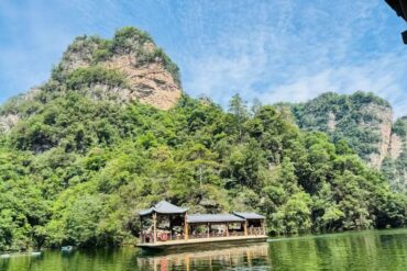 Baofeng Lake,Zhangjiajie,Chinatour Wooden pavilion-style boat on a calm green river, with forested cliffs and a blue sky above.