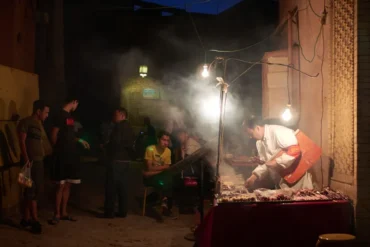 Kuanzhai Alleys,Chengdu,Chinatour Night street food stall with a cook grilling skewers under hanging bulbs as customers wait nearby in a smoky, dimly lit alley.