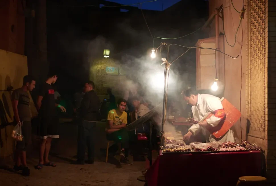 Kuanzhai Alleys,Chengdu,Chinatour Night street food stall with a cook grilling skewers under hanging bulbs as customers wait nearby in a smoky, dimly lit alley.