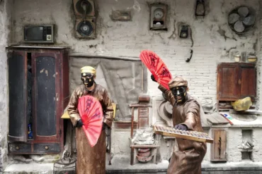 Two masked performers in brown robes wield red folding fans in a dusty, rustic set with old cupboards and vintage appliances on the wall.