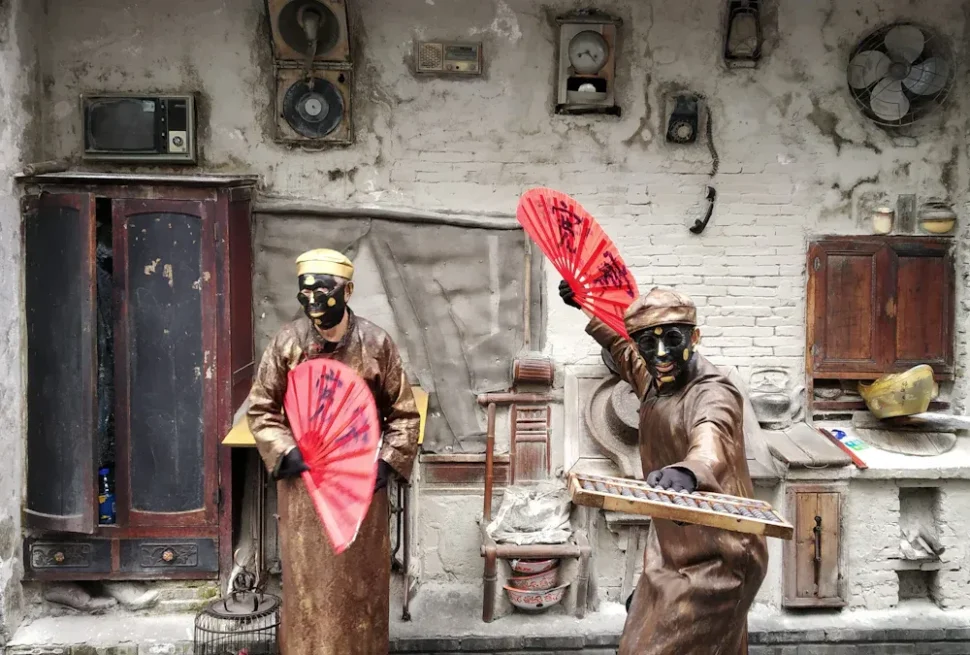 Two masked performers in brown robes wield red folding fans in a dusty, rustic set with old cupboards and vintage appliances on the wall.