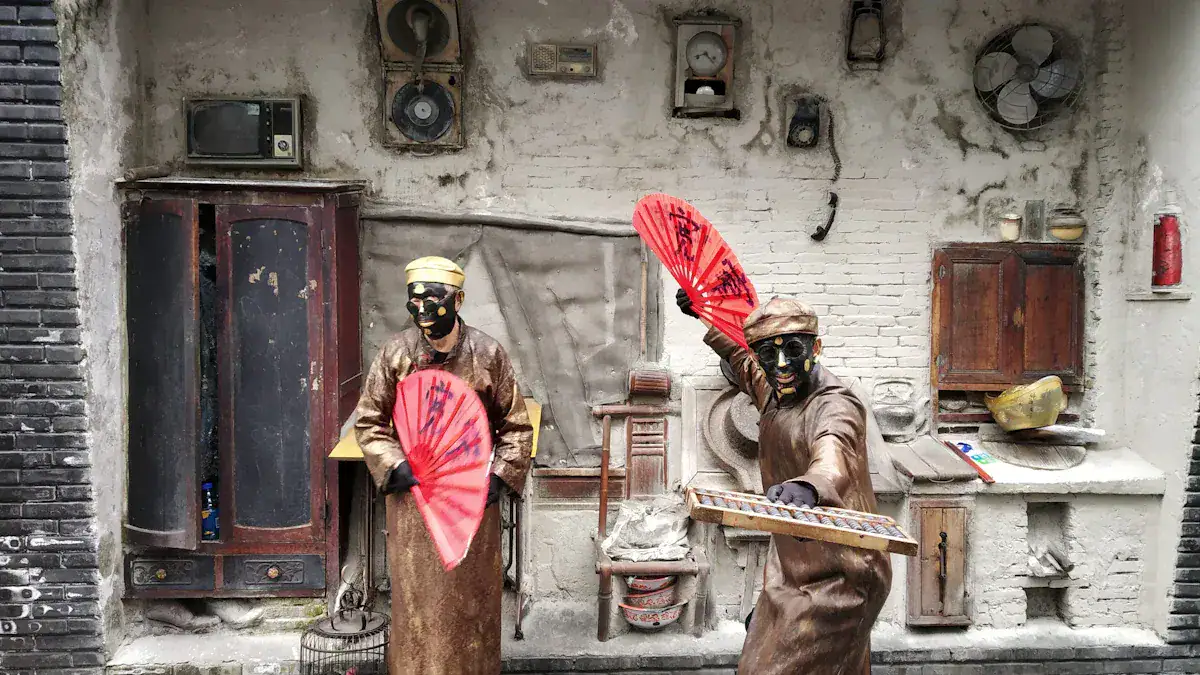 Two masked performers in brown robes wield red folding fans in a dusty, rustic set with old cupboards and vintage appliances on the wall.