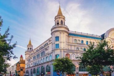 Historic European-style shopping arcade with two tower spires and arched storefronts along a tree-lined street on a sunny day.