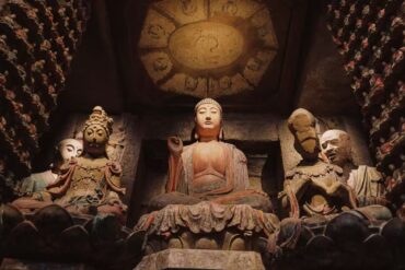 Central Buddha statue seated in a cave temple with smaller statues around, dimly lit and ancient stone carvings behind it