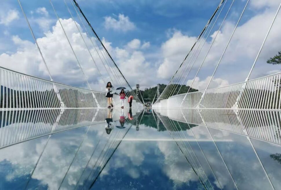 Tianmen Mountain Cableway and Glass Skywalk,Zhangjiajie,Chinatour People walk on a glass-bottom bridge with a clear blue sky, their reflections visible in the mirrored surface below.