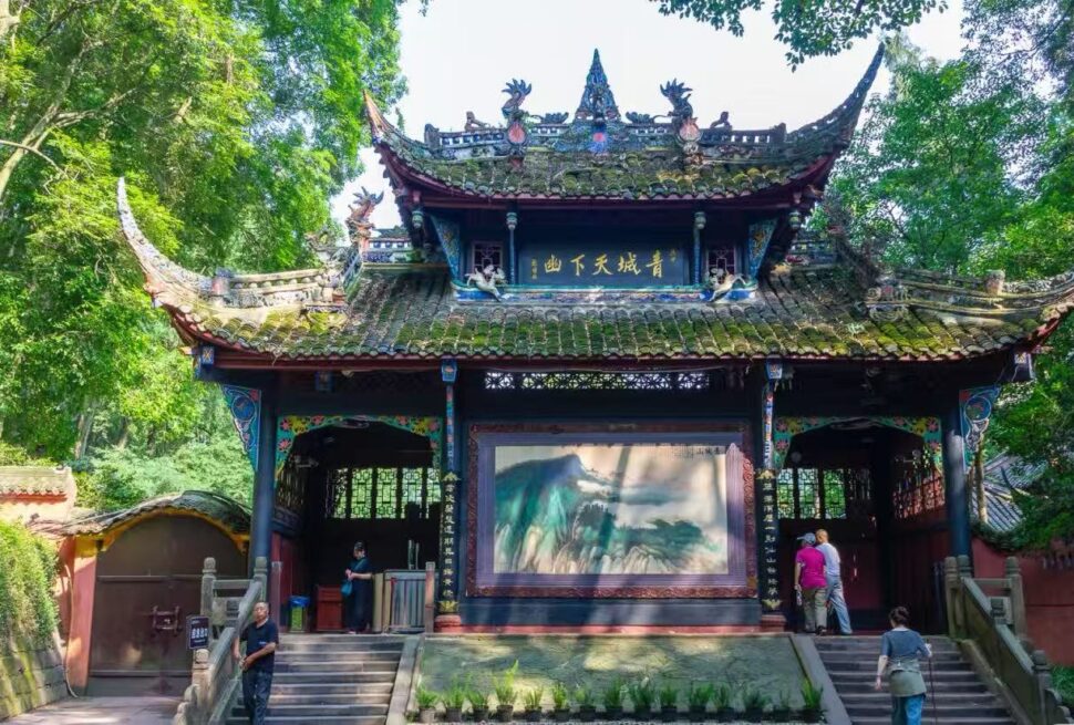 Traditional Chinese pavilion tucked in a lush green garden with ornate roof dragons and a large landscape painting displayed inside the open hall, visitors examining the artwork.