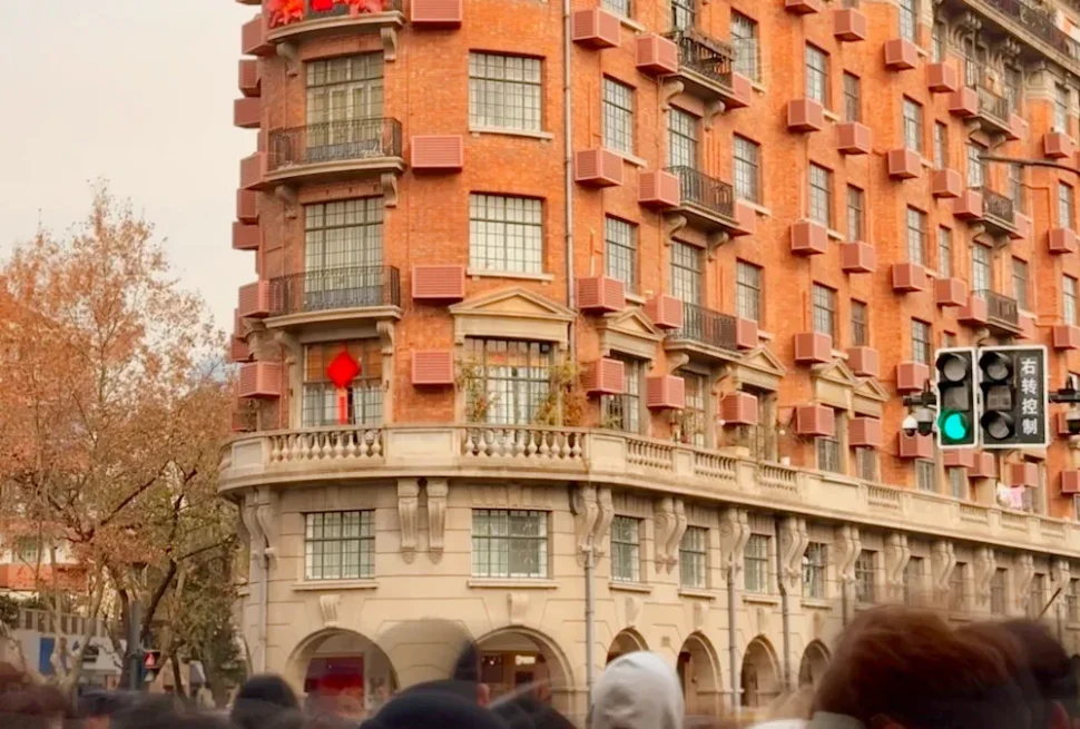 Curved red-brick building with many protruding balconies and arched ground floors on a busy city street, pedestrians in foreground.
