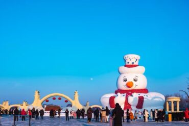 Large inflatable snowman sculpture with red scarf in an icy park, crowded with people under a clear blue sky at a winter festival.