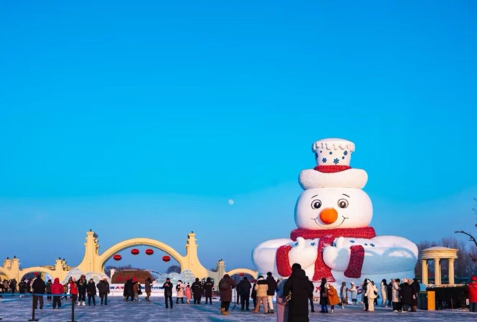 Large inflatable snowman sculpture with red scarf in an icy park, crowded with people under a clear blue sky at a winter festival.
