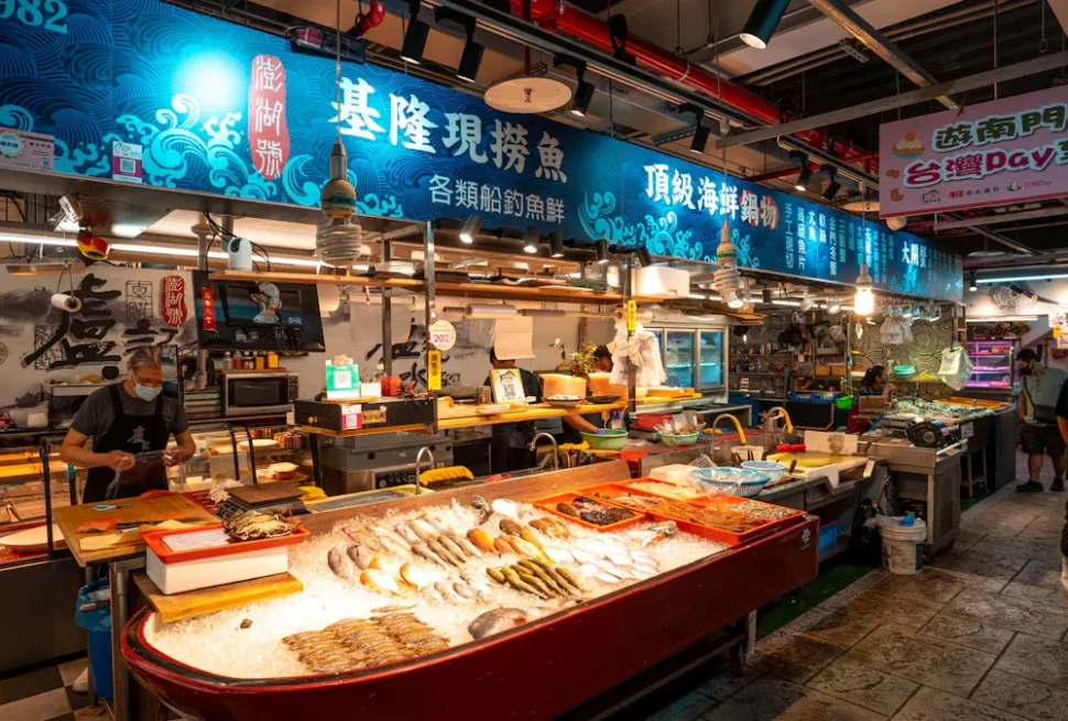Seafood market stall with an ice-filled display of fish and shellfish, blue decorative signs overhead, vendors at work in the background.