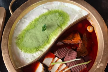 Split hot pot with milky white broth topped with green powder on the left and a red spicy broth with vegetables on the right.