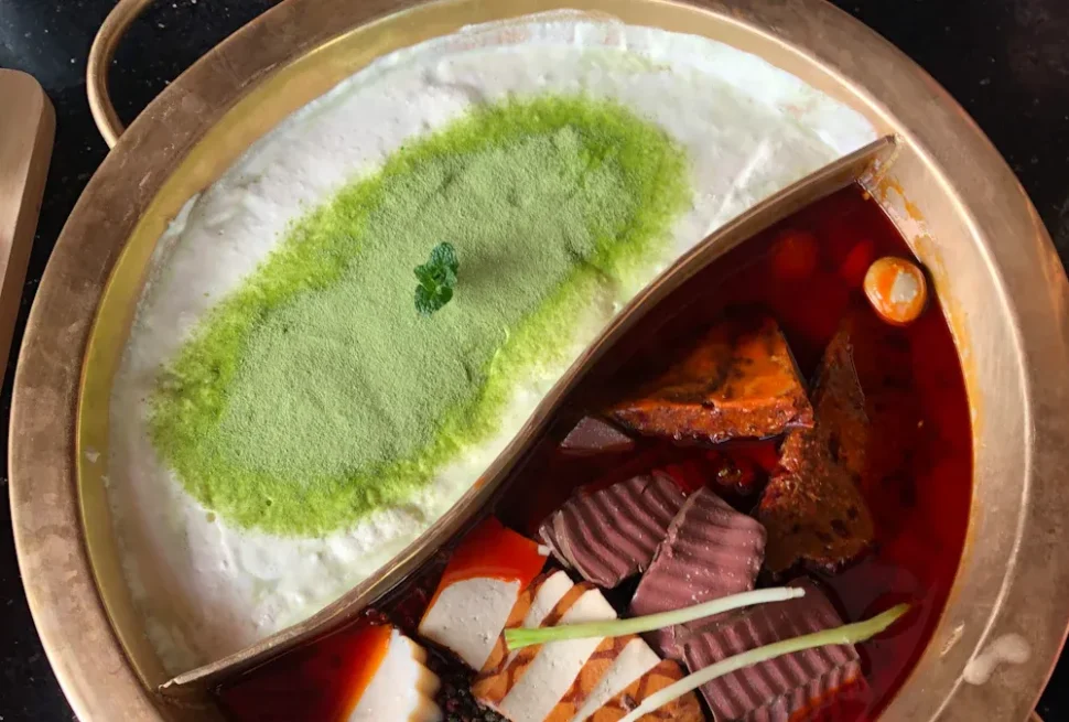 Split hot pot with milky white broth topped with green powder on the left and a red spicy broth with vegetables on the right.