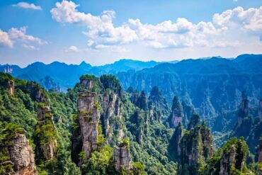 Towering sandstone pinnacles covered in green trees rise in a mountainous valley under a blue sky with fluffy clouds.