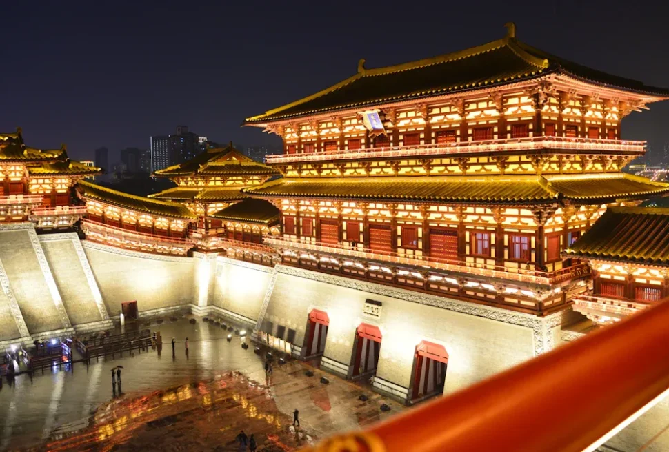 Nighttime view of a large illuminated Chinese palace complex with multi-tiered roofs and red walls, set in a spacious courtyard area.