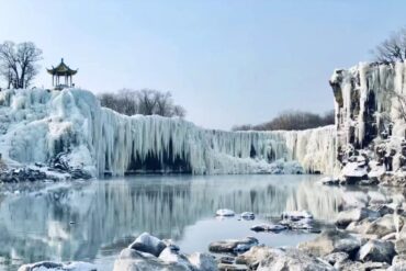 Winter scene with a frozen waterfall and icicles, a small pagoda on the left, and a calm, reflective lake below.