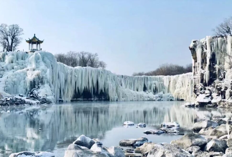 Jingpo Lake and Diaoshuilou Waterfall,Mudanjiang,Chinatour Winter scene with a frozen waterfall and icicles, a small pagoda on the left, and a calm, reflective lake below.