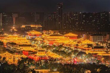Nighttime panorama of a brightly lit traditional Chinese palace complex with glowing orange rooftops against a modern city skyline.