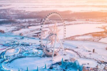 Aerial view of a large, illuminated Ferris wheel in a snow-covered amusement park at sunset, with crowds below.