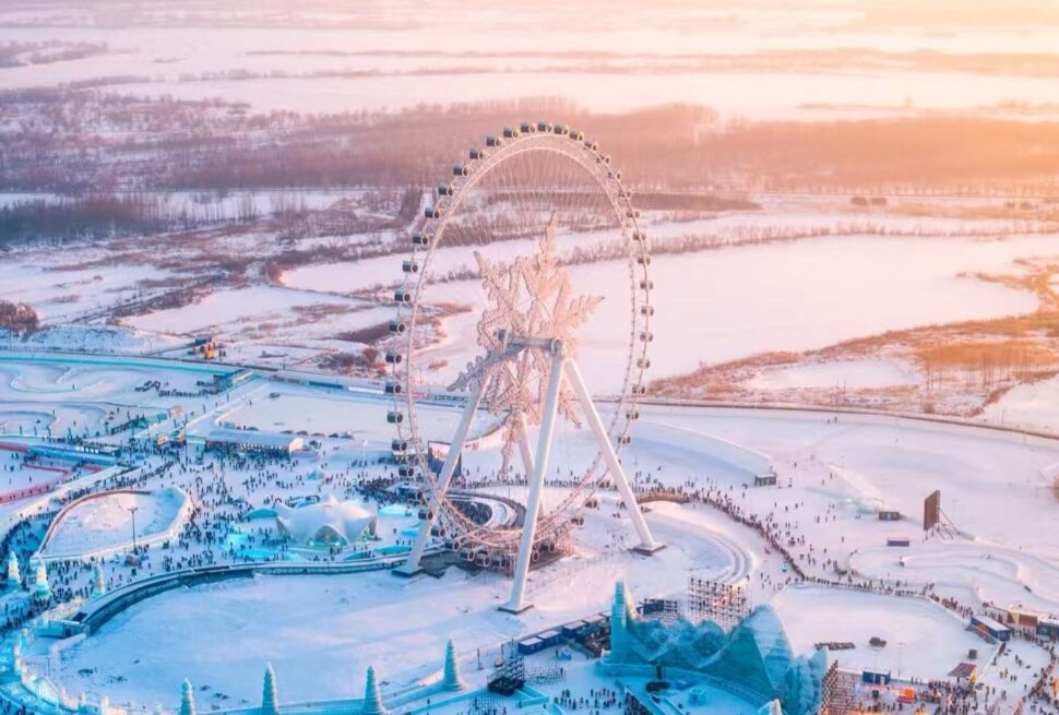 Aerial view of a large, illuminated Ferris wheel in a snow-covered amusement park at sunset, with crowds below.