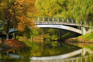 Bridge spans a calm river in a park, autumn leaves in orange and yellow on trees, green willows nearby, reflections in the water.