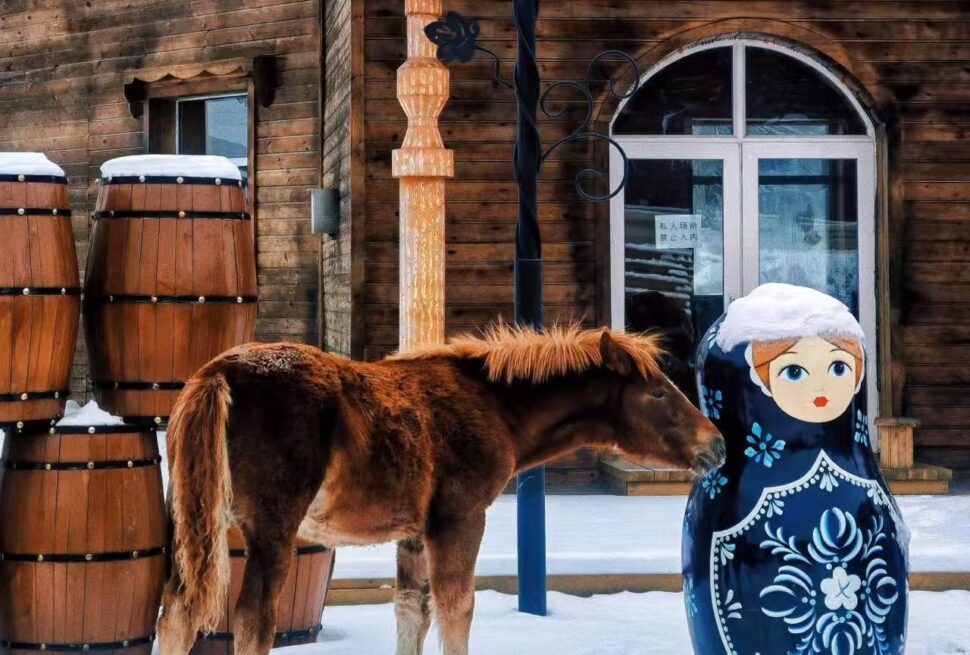 Brown horse sniffs a large blue Matryoshka-style doll in a snowy yard beside wooden barrels and a cabin.