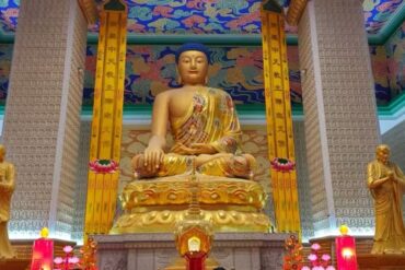 Golden seated Buddha statue in a richly decorated temple hall, flanked by two smaller statues, with a colorful ornate ceiling and illuminated altar candles and flowers behind it.