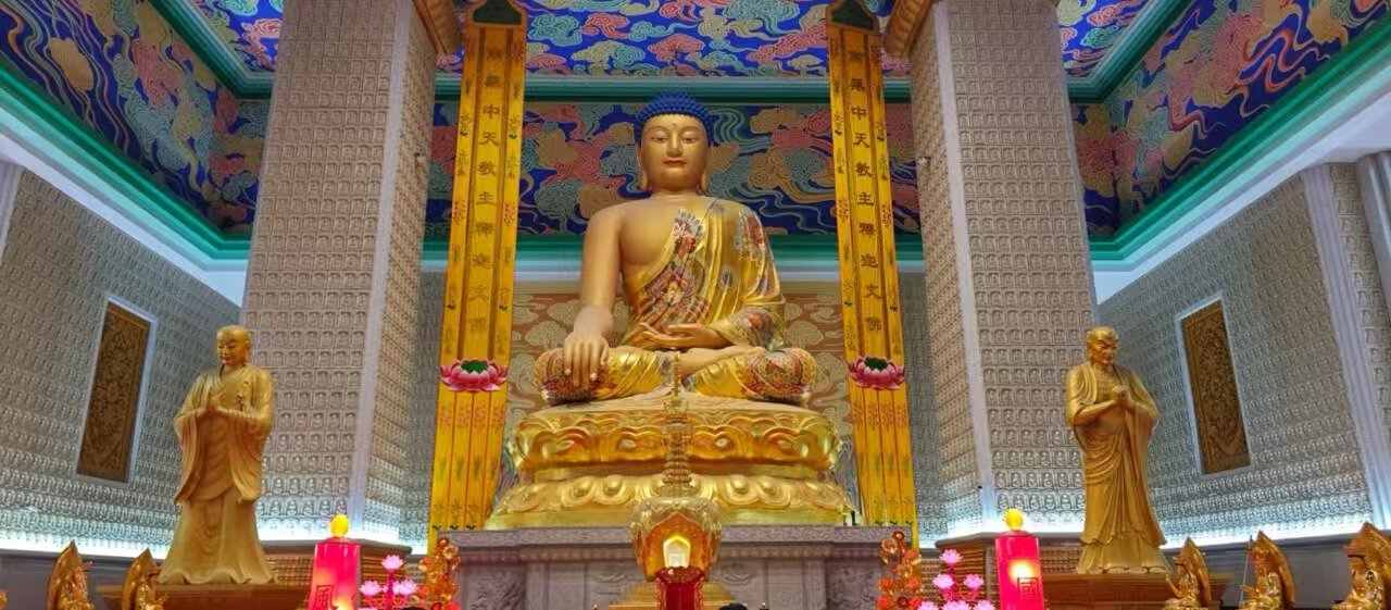 Famen Temple,X`an,Chinatour Golden seated Buddha statue in a richly decorated temple hall, flanked by two smaller statues, with a colorful ornate ceiling and illuminated altar candles and flowers behind it.