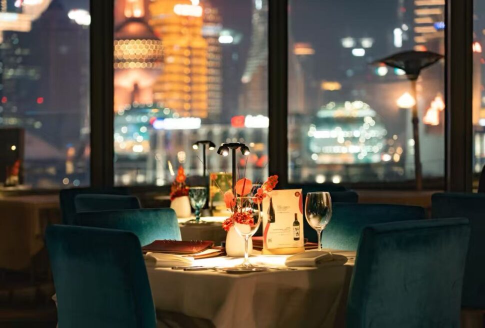 Table for two in a dim restaurant with teal chairs, a floral centerpiece, wine glass, and city lights outside the windows.